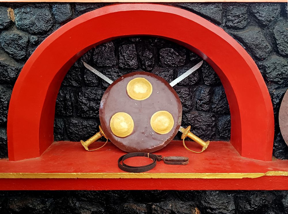 Shield and crossed swords set within the red arch of the Kalari wall